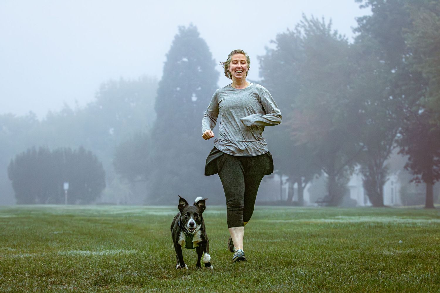 Smiling woman jogging with a Boston Terrier on a foggy green park lawn.