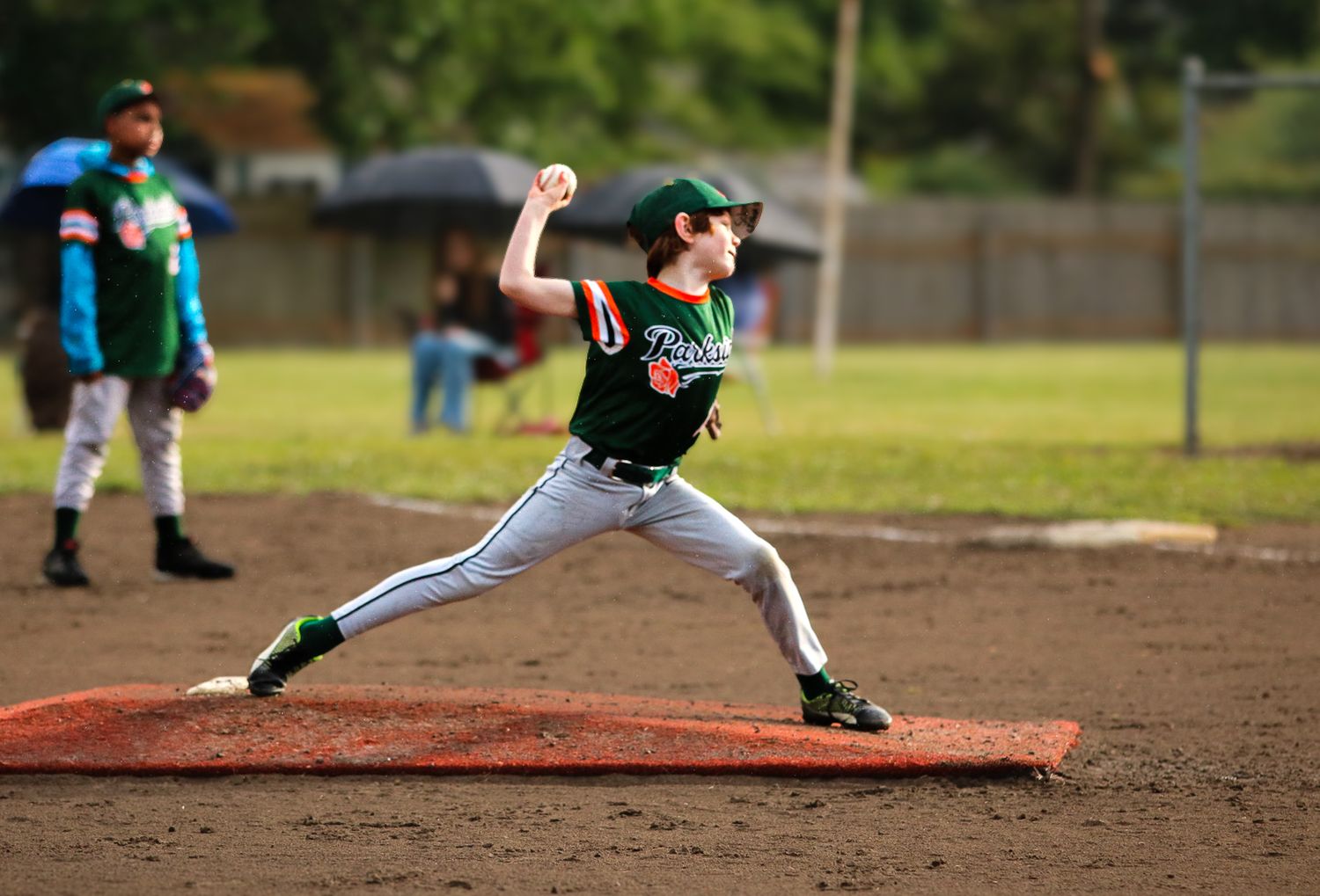 Young baseball pitcher in green uniform winding up to throw on a youth league field.