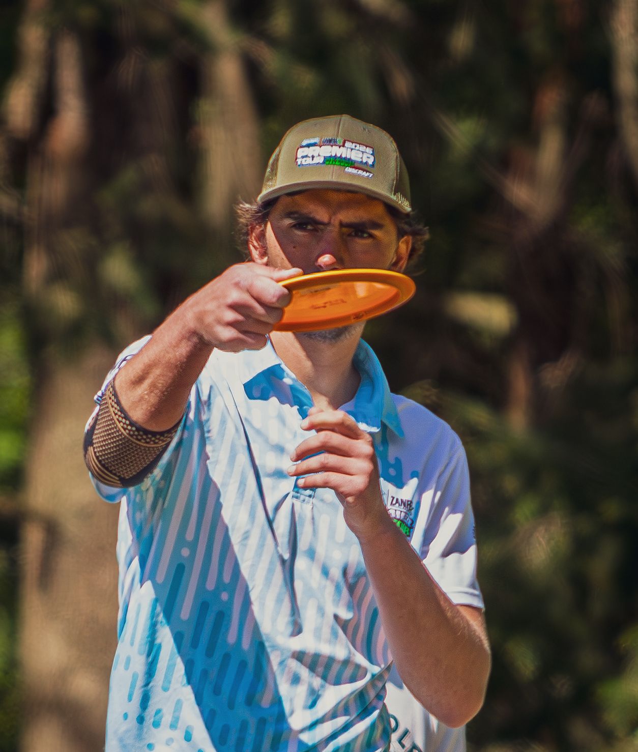 Male disc golf player holding an orange disc on a sunny wooded course.