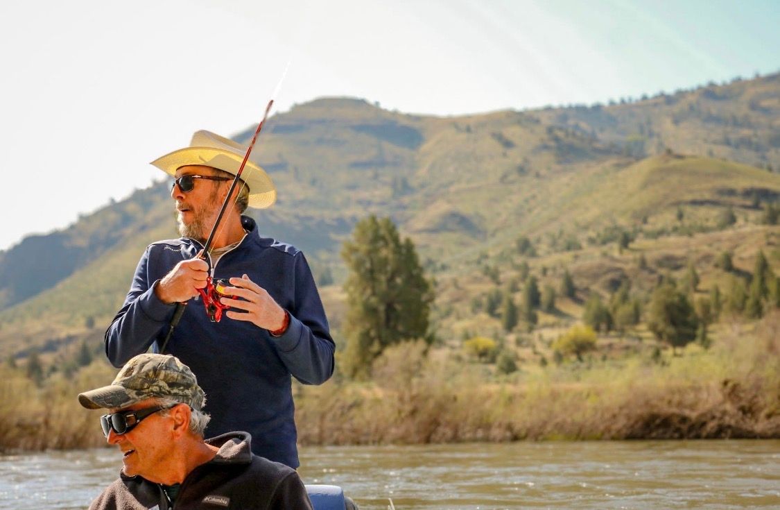 A man in a straw hat holds a fishing rod while seated in a boat on a mountain river.
