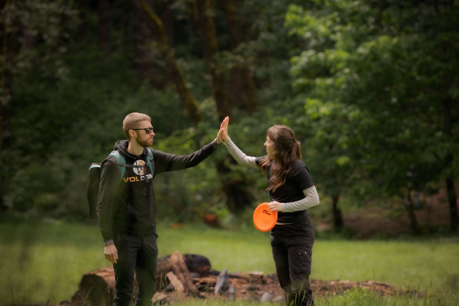 Two people high-fiving in a forest clearing while one holds an orange disc golf frisbee.