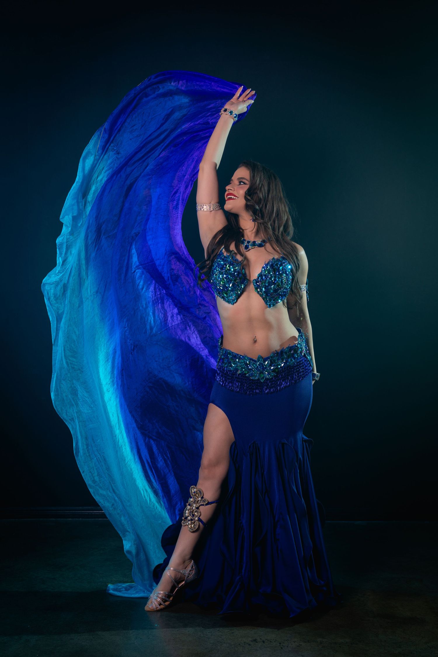 A belly dancer in a blue sequined costume dramatically waves flowing blue silk veils against a dark background.