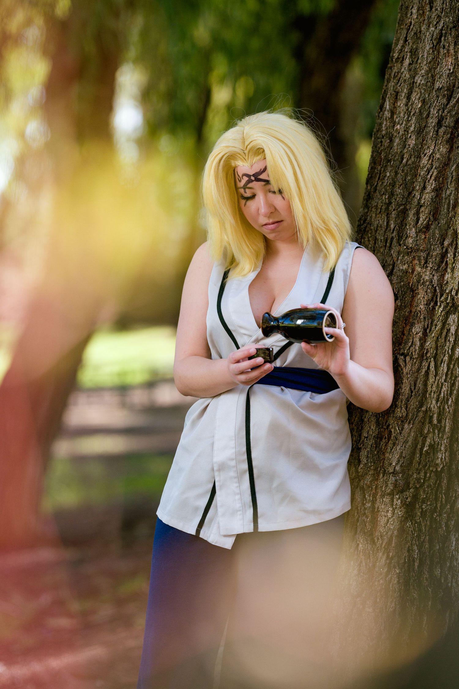 Blonde woman cosplaying as Tsunade from Naruto leaning against a tree in a sunny park setting.