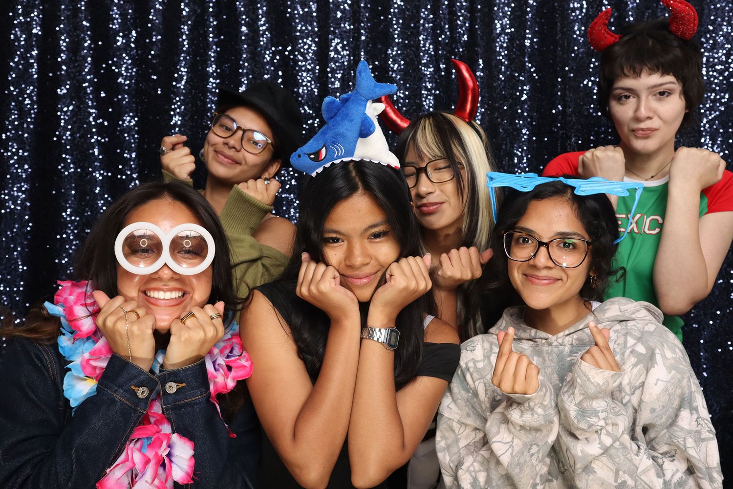 Five young women pose playfully in a photo booth with fun props including glasses, devil horns, and a shark hat.