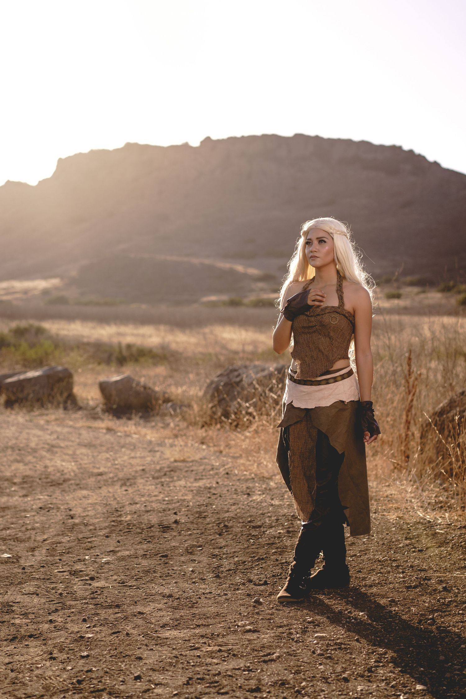 Blonde woman in fantasy warrior costume standing in a sunlit desert landscape with rocky cliffs.