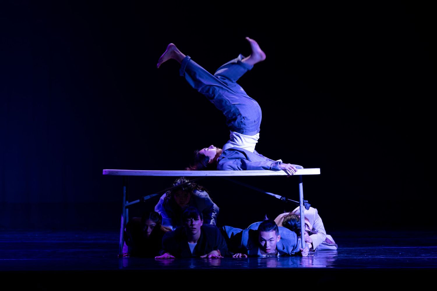 Acrobat balances headstand on table while performers pose dramatically beneath in blue-lit stage act.
