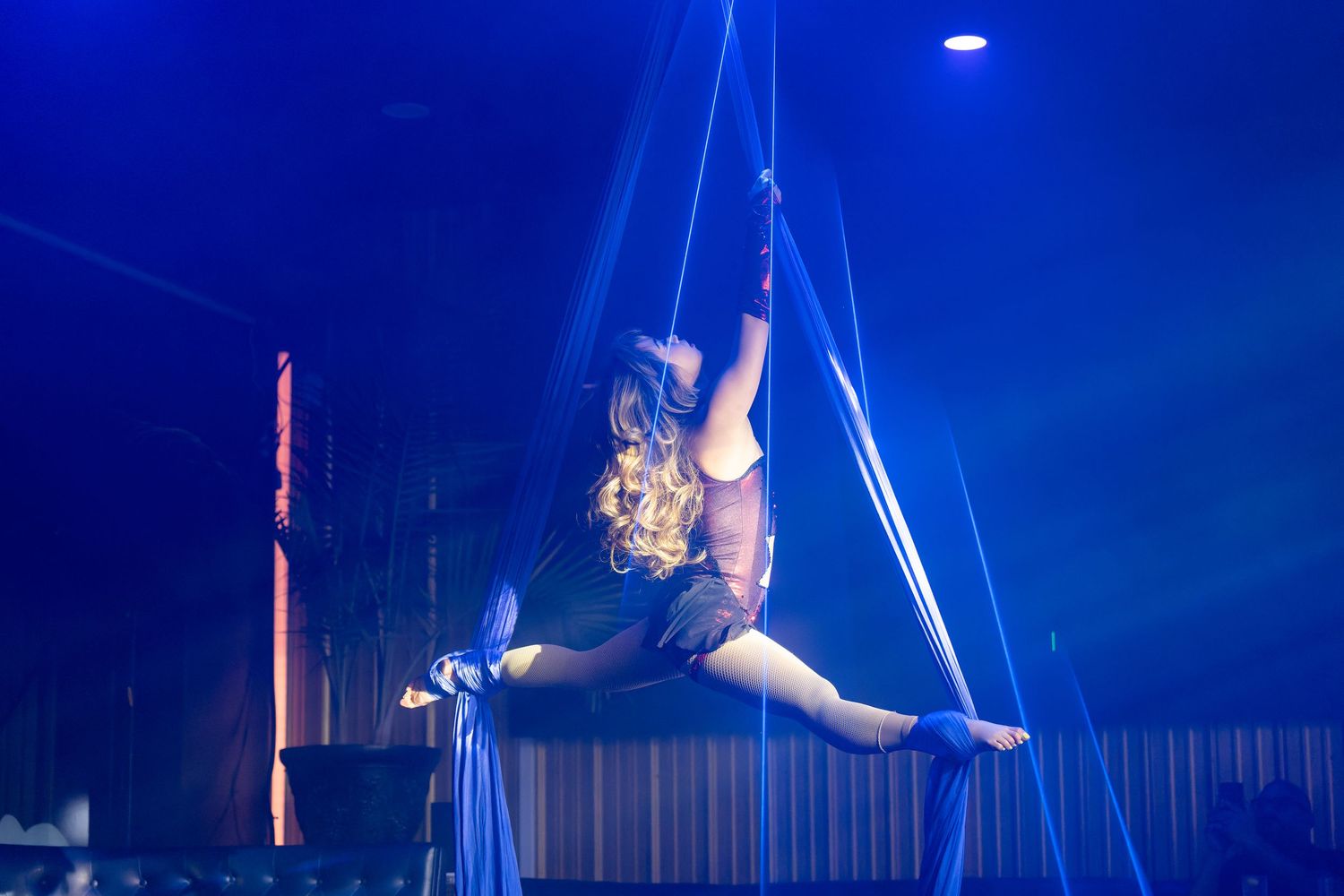 Aerial acrobat performs stunning splits on hanging silks under blue stage lighting at a circus performance.