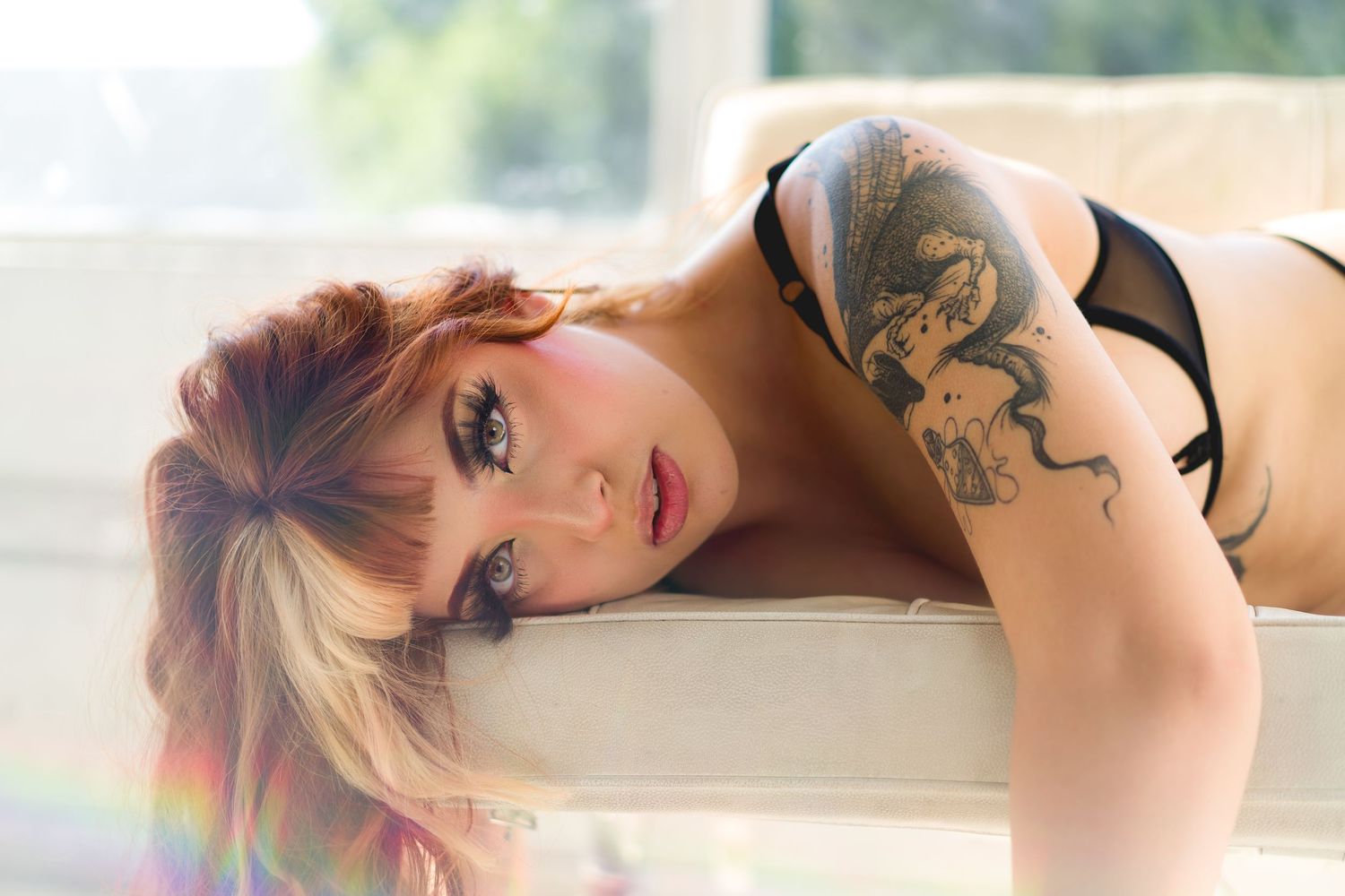 Woman with snake tattoo on arm poses in black lingerie on white furniture.
