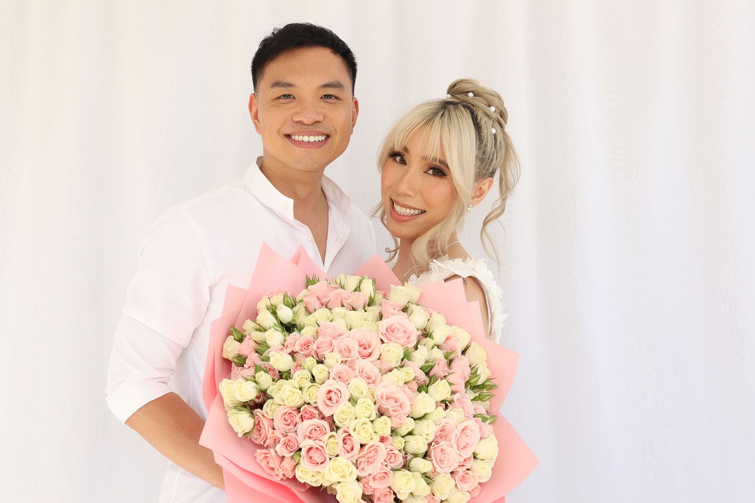 Smiling couple posing together holding a large bouquet of pink roses against a white background.