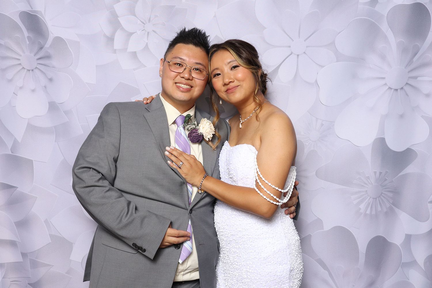 Couple posing at wedding photo booth with floral backdrop, groom in gray suit and bride in white dress.