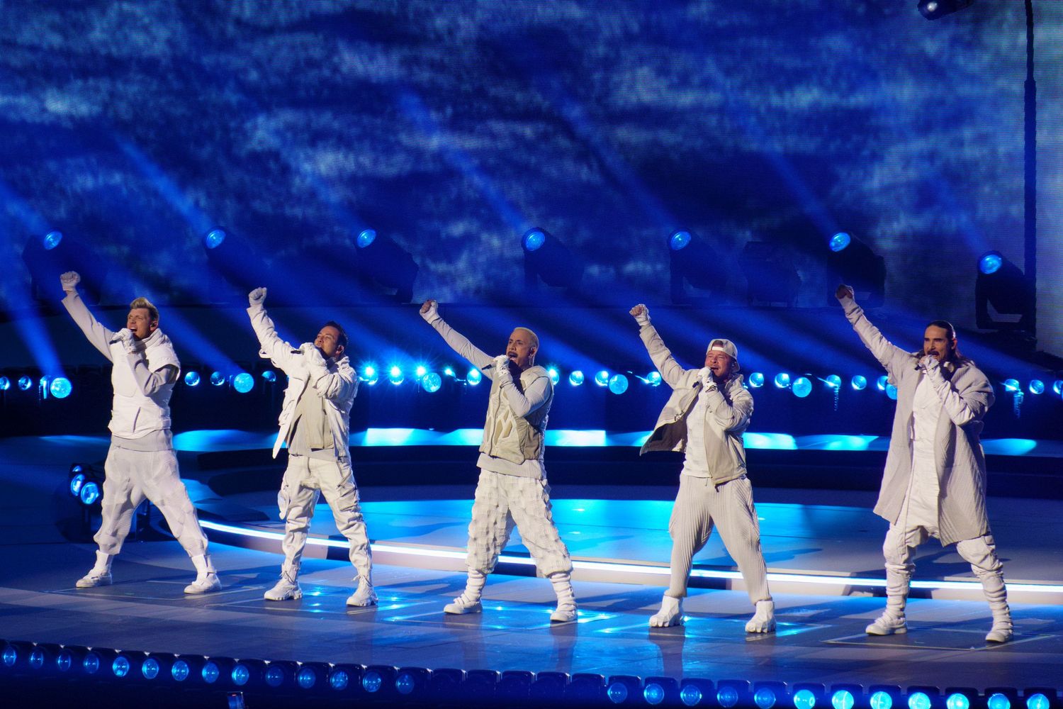 Five male performers in matching white outfits dance in sync on a large lit stage.