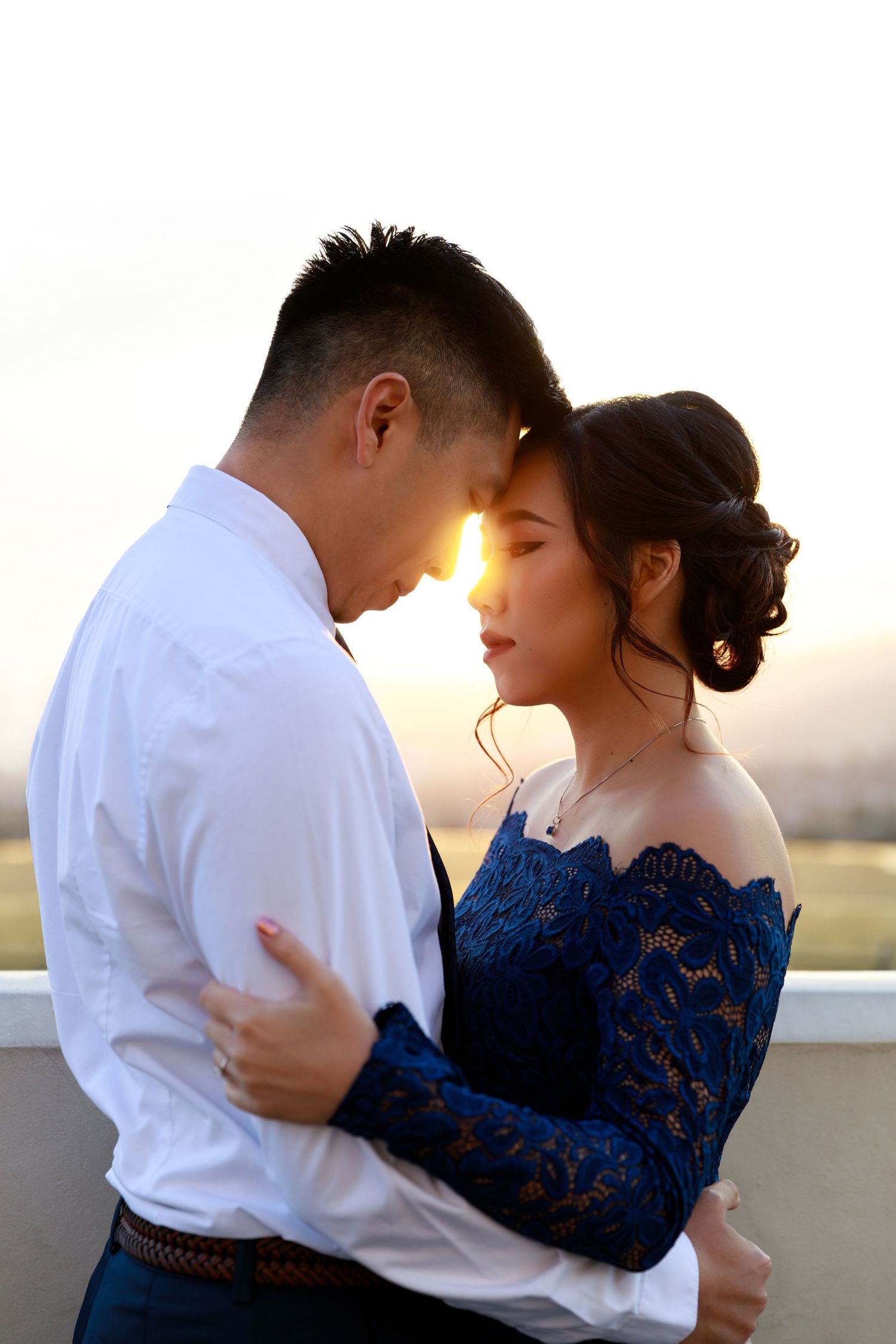 A couple embraces tenderly at sunset, foreheads touching, woman in blue lace off-shoulder dress and man in white shirt.