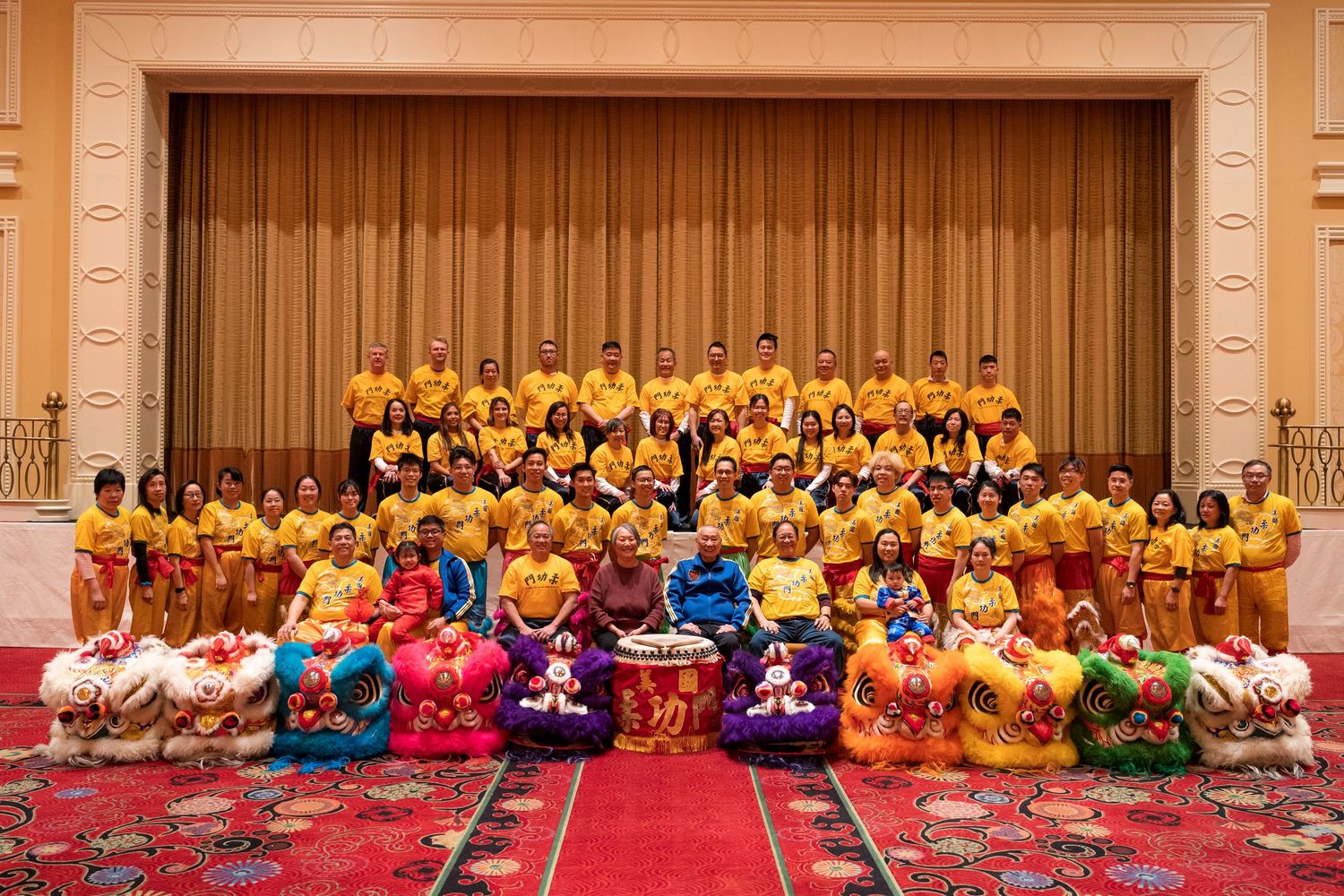 Large group in yellow traditional costumes poses with colorful lion dance props on red carpet.