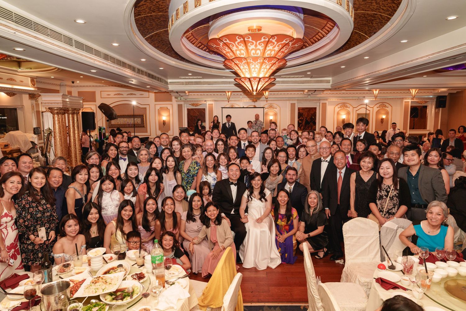 Large wedding group photo with bride and groom surrounded by guests in an elegant banquet hall.
