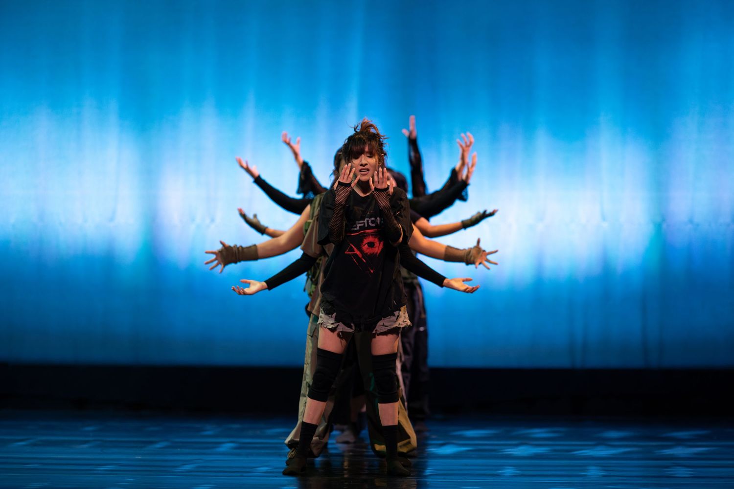 Group of dancers create multi-armed Kali goddess illusion on blue-lit stage in striking performance art.