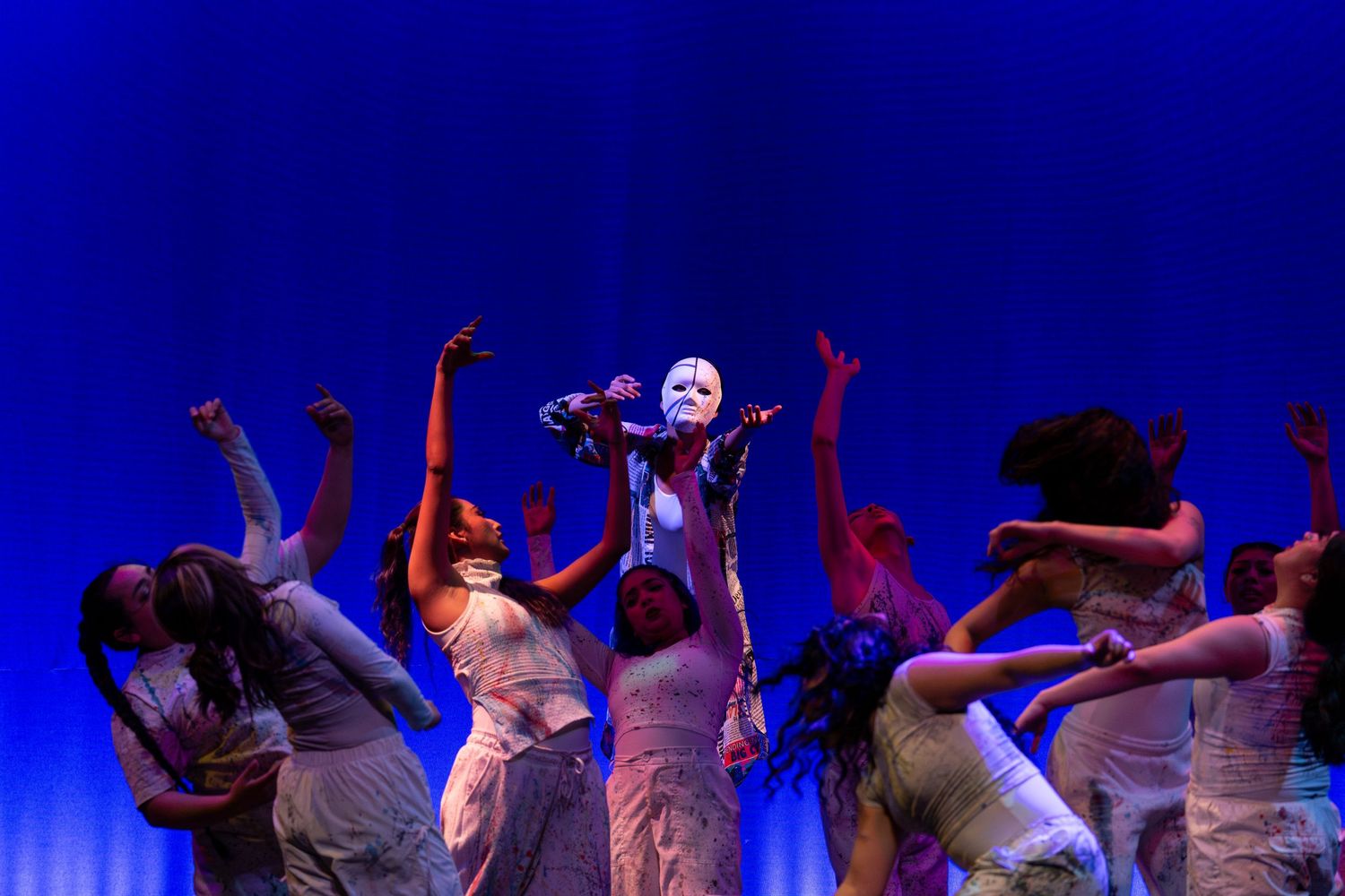 Masked performer leads ensemble of white-clad dancers under vivid blue stage lighting in theatrical show.