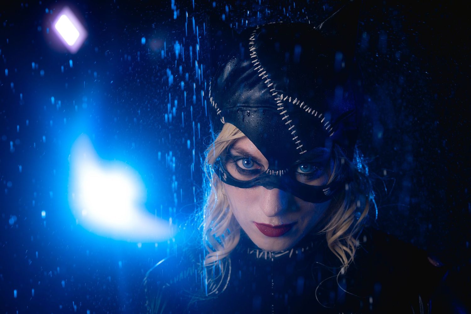 Blonde woman in Catwoman mask and costume in dramatic blue rainy night scene.