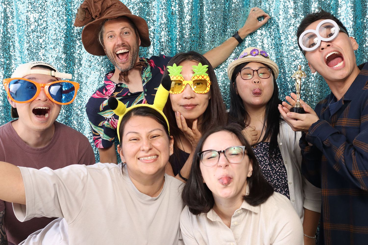 Group of seven friends posing with fun novelty glasses and props in front of a teal sequin backdrop at a photo booth.