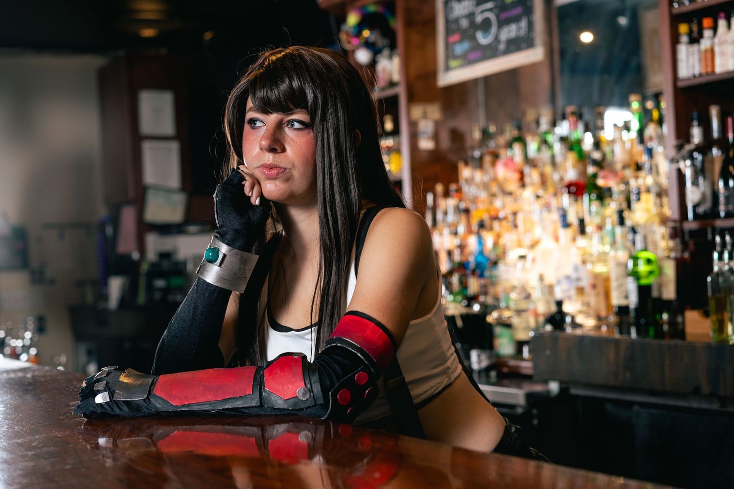 A woman in cosplay attire with red and black gloves leans on a bar counter with a thoughtful expression.