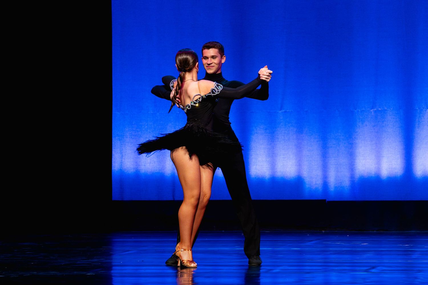 Elegant couple performs Latin ballroom dance in matching black attire against a blue stage backdrop.