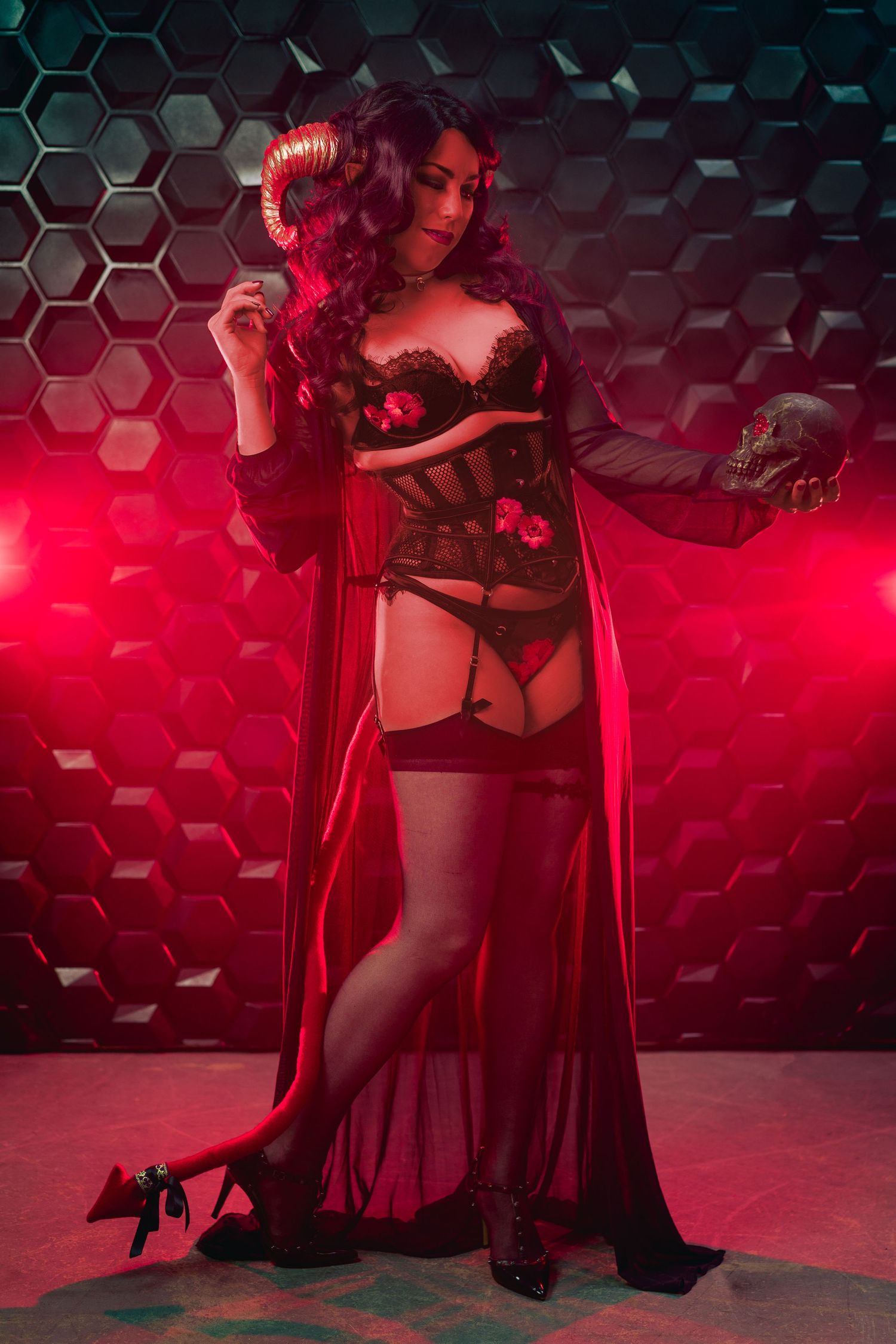 Woman in devil cosplay with horns wearing black lingerie and red robe holding a skull in red lighting.