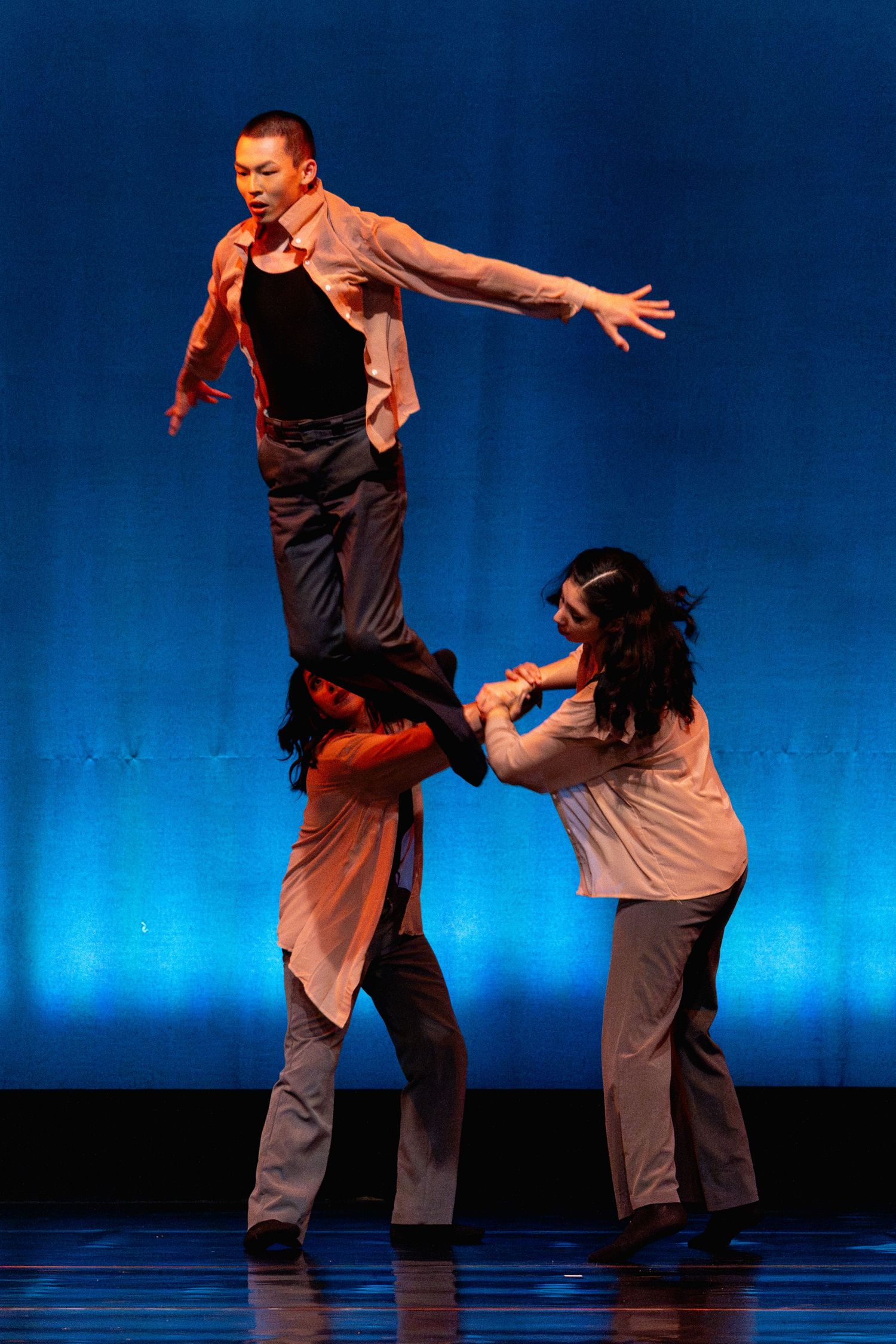 Two dancers perform impressive acrobatic lift with male dancer balanced overhead on blue-lit stage.