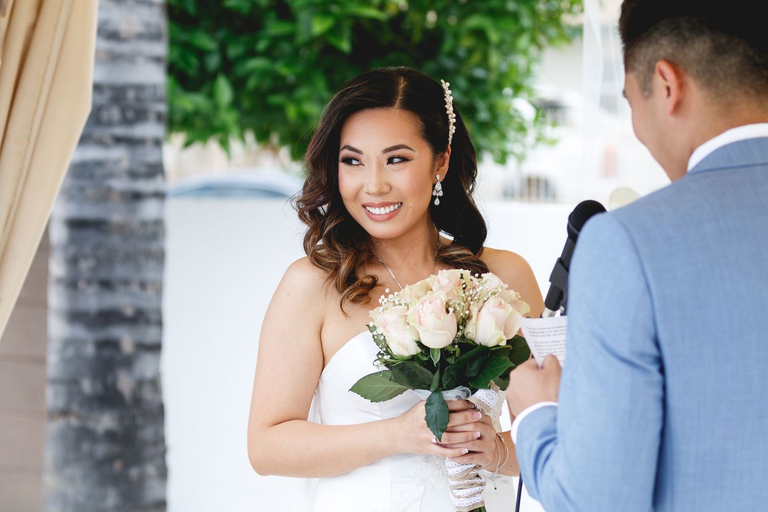 Smiling Asian bride holds white rose bouquet during outdoor wedding ceremony.
