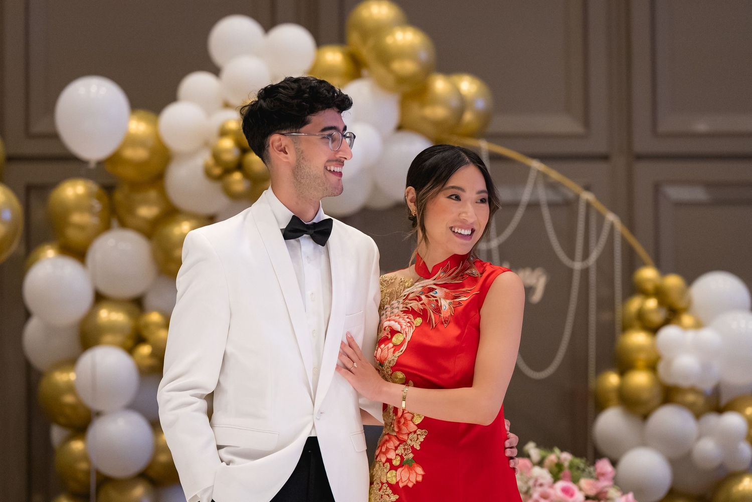 Couple poses in front of gold and white balloon arch, groom in white tuxedo, bride in red qipao.