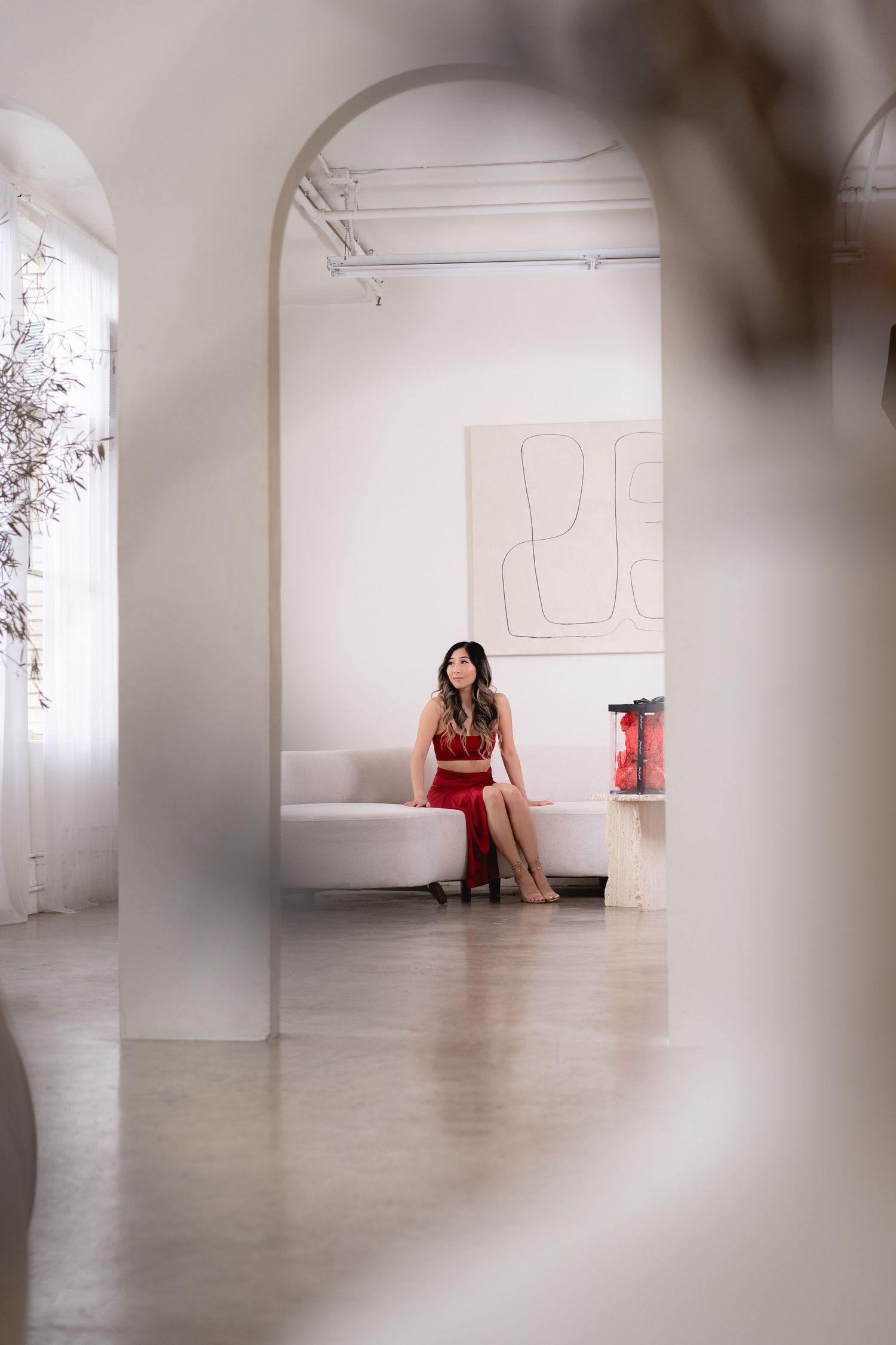 Woman in red two-piece outfit sits on white sofa in minimalist white studio.