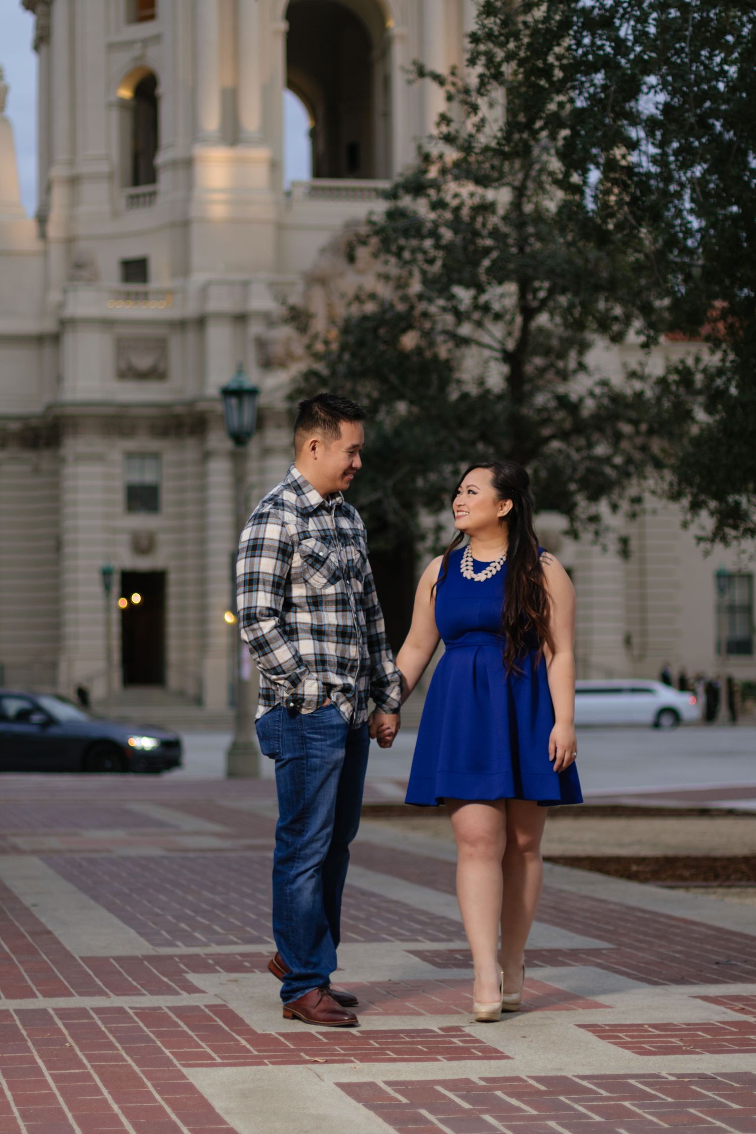 Couple holds hands and gazes at each other near grand building at dusk.