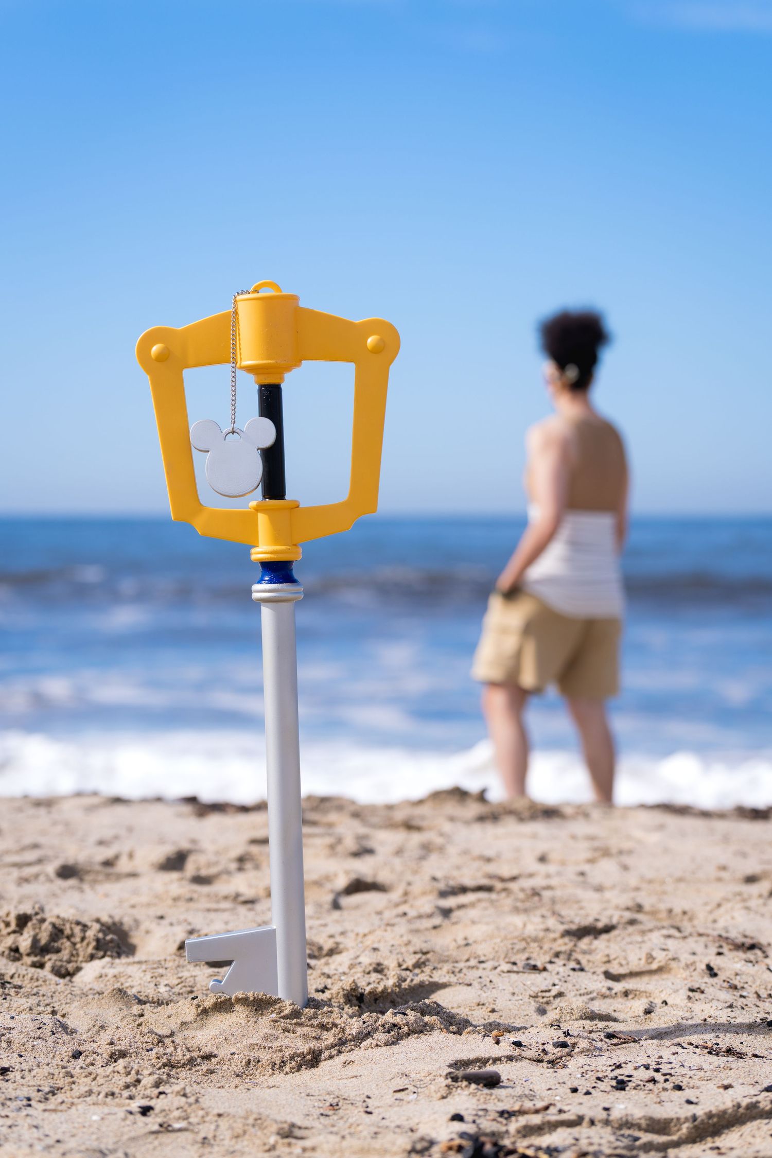 Yellow Kingdom Hearts Keyblade prop standing in beach sand with person looking at ocean in background.