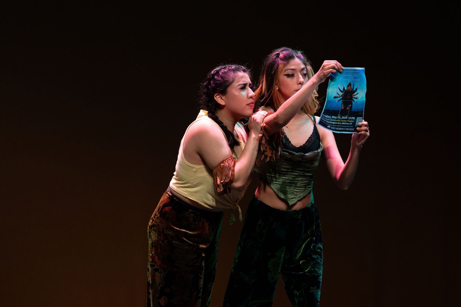 Two young female performers examine a book together under warm amber stage lighting in theater.