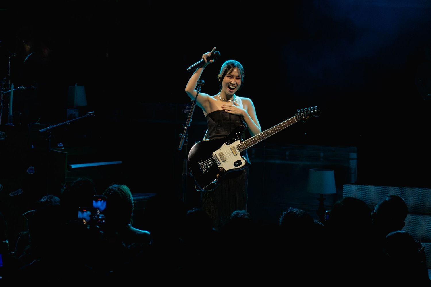 Female guitarist performs energetically on a dark concert stage with electric guitar.