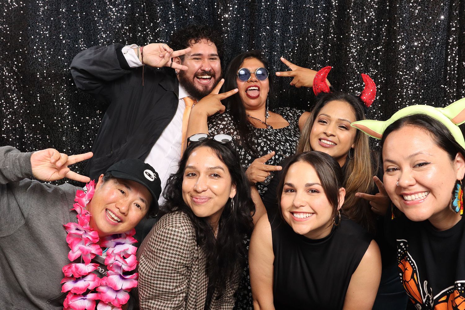 Group of eight friends posing with fun accessories in a photo booth with a black sparkle backdrop.
