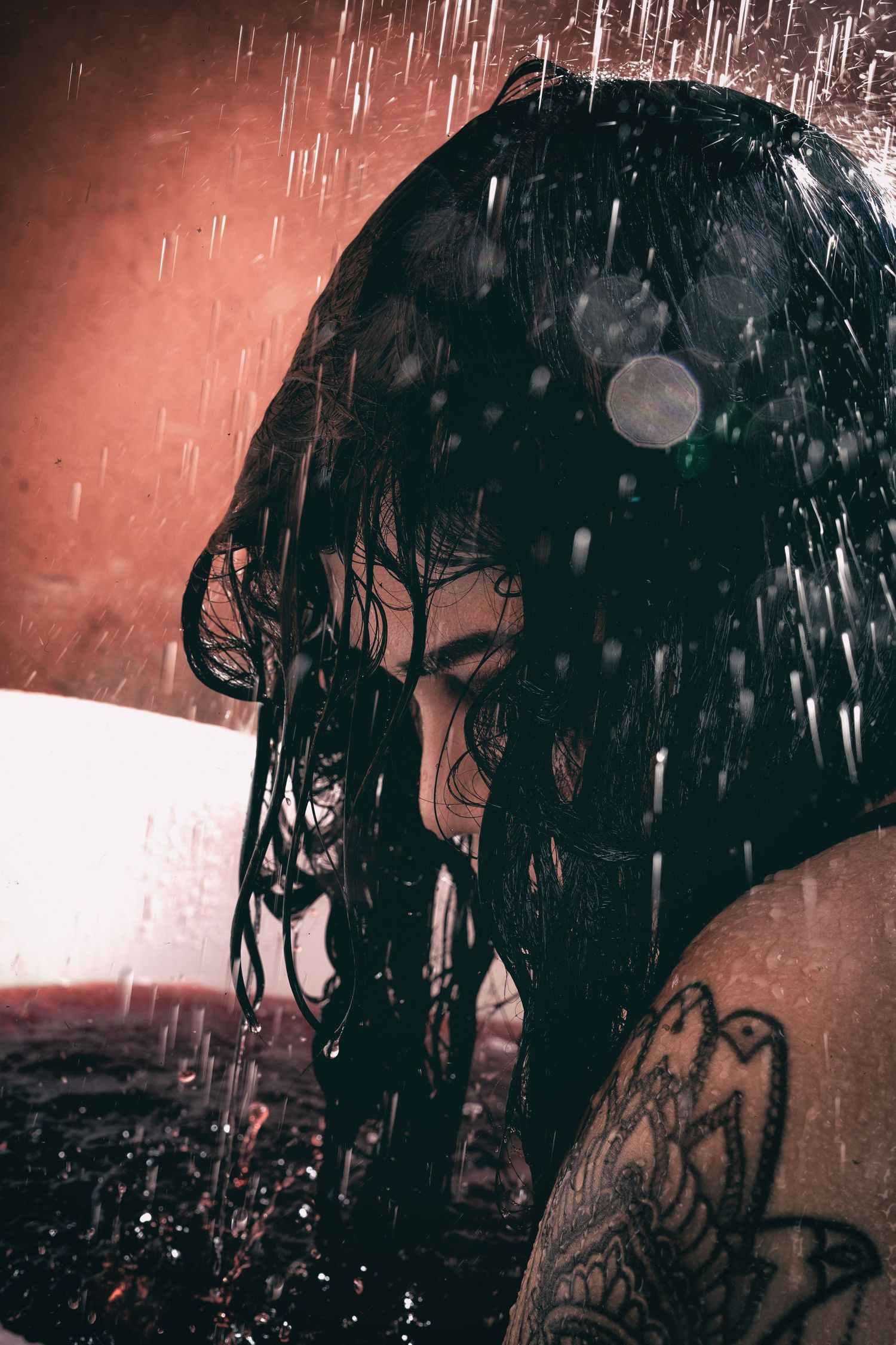 Woman with wet dark hair and tattoo standing in shower with water droplets falling.