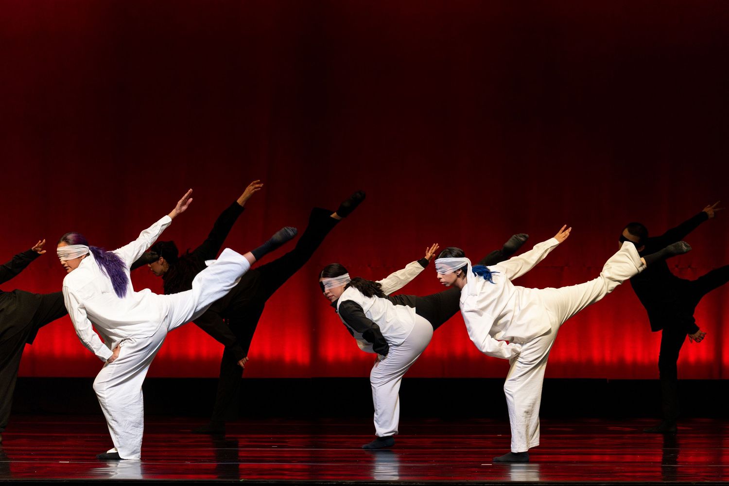 Synchronized dancers in white uniforms execute high kicks against dramatic red backdrop on stage.