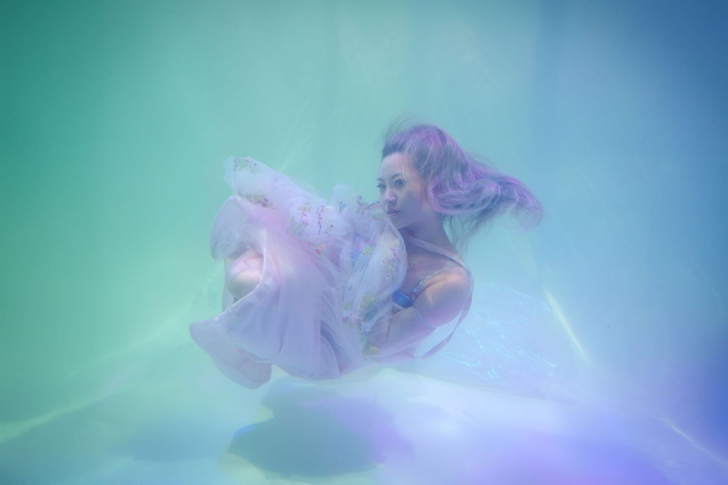 Woman in purple dress floating underwater in turquoise and purple illuminated pool.