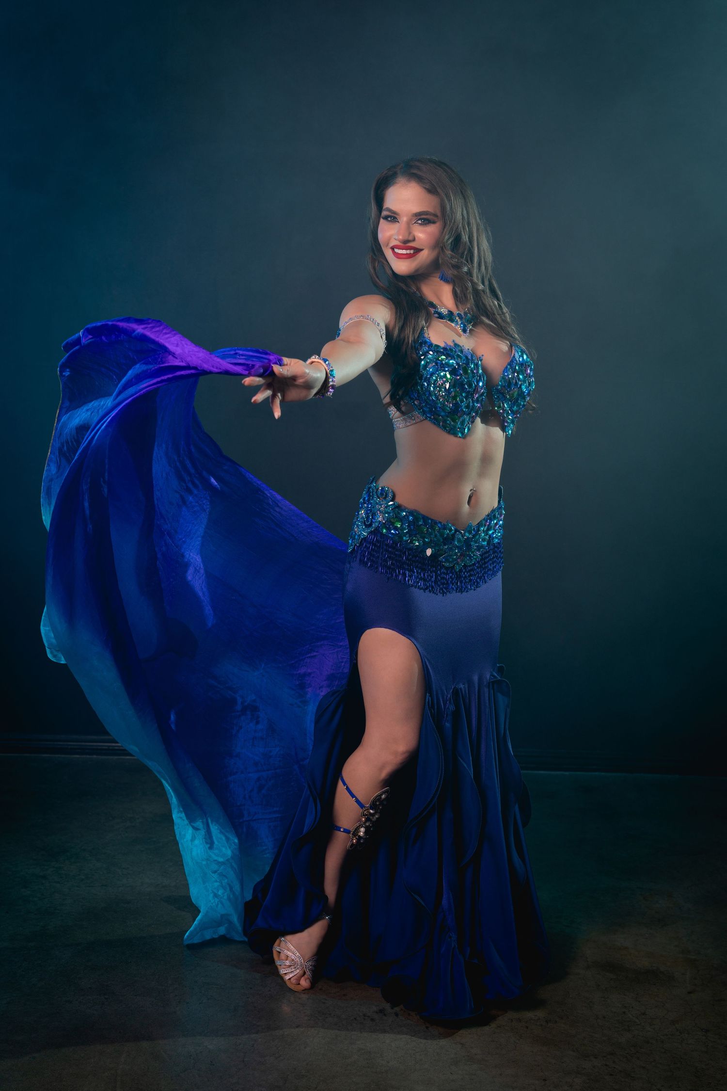 Smiling woman in blue sequined belly dance costume performing with flowing silk veil.