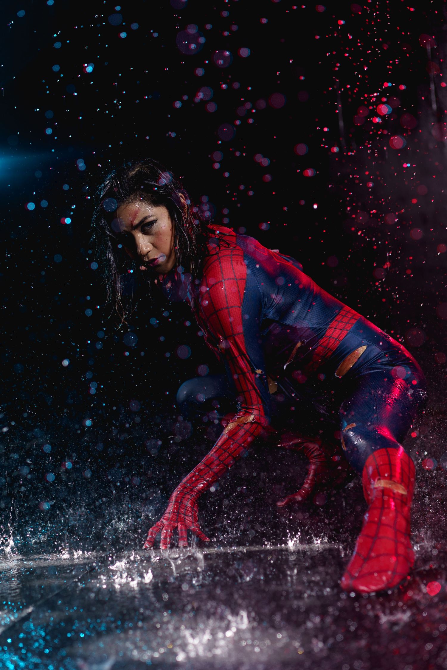 Woman in Spider-Man cosplay crouching in dynamic pose with colorful water splashes on dark background.