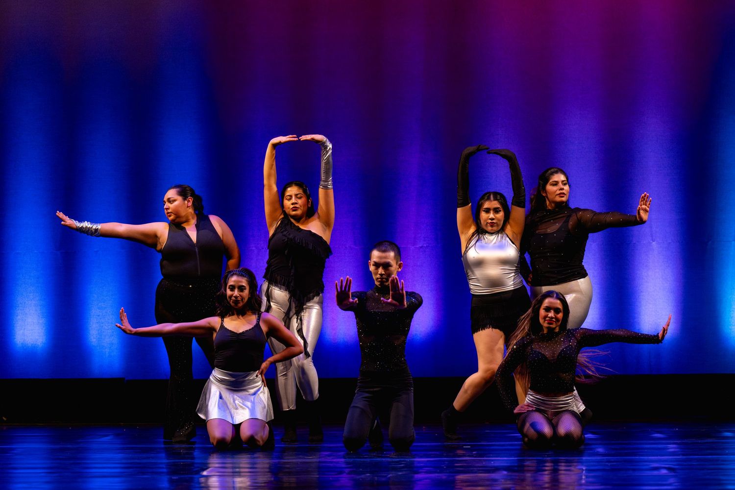 Seven dancers perform on a dramatic blue and purple lit stage in black and white costumes.