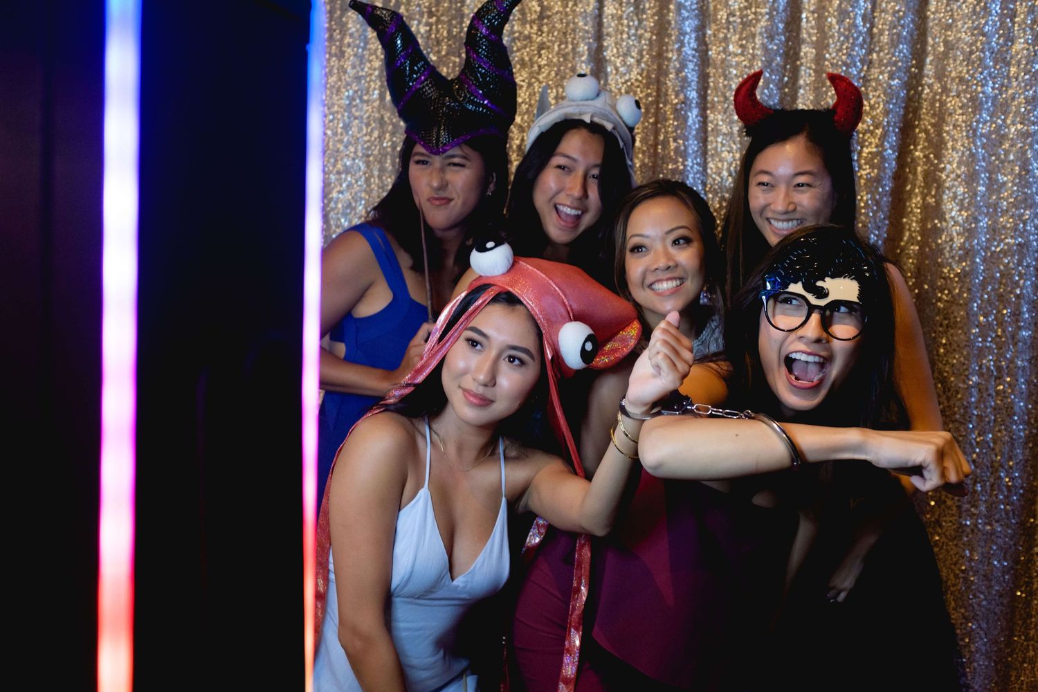 Six young women pose playfully in a photo booth wearing fun party props including devil horns and bunny ears.