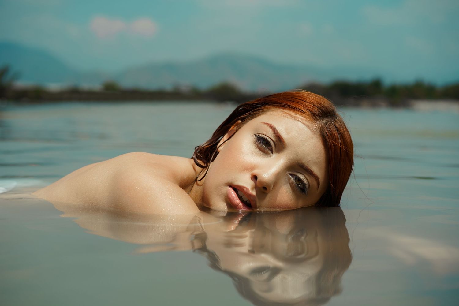 Redhead woman lying in shallow water with mountains visible in the background under cloudy skies.