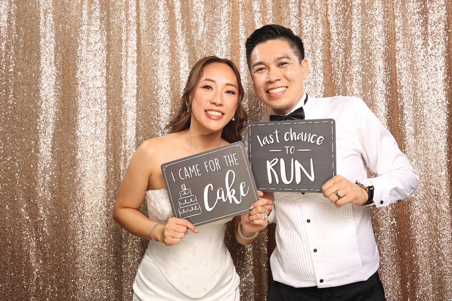 Couple at wedding photo booth holding funny signs about cake and last chance to run, posing in front of gold sequin backdrop.