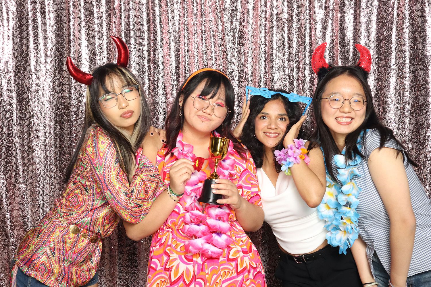 Four young women posing in a photo booth with devil horns headbands and floral leis against a silver sequin backdrop.