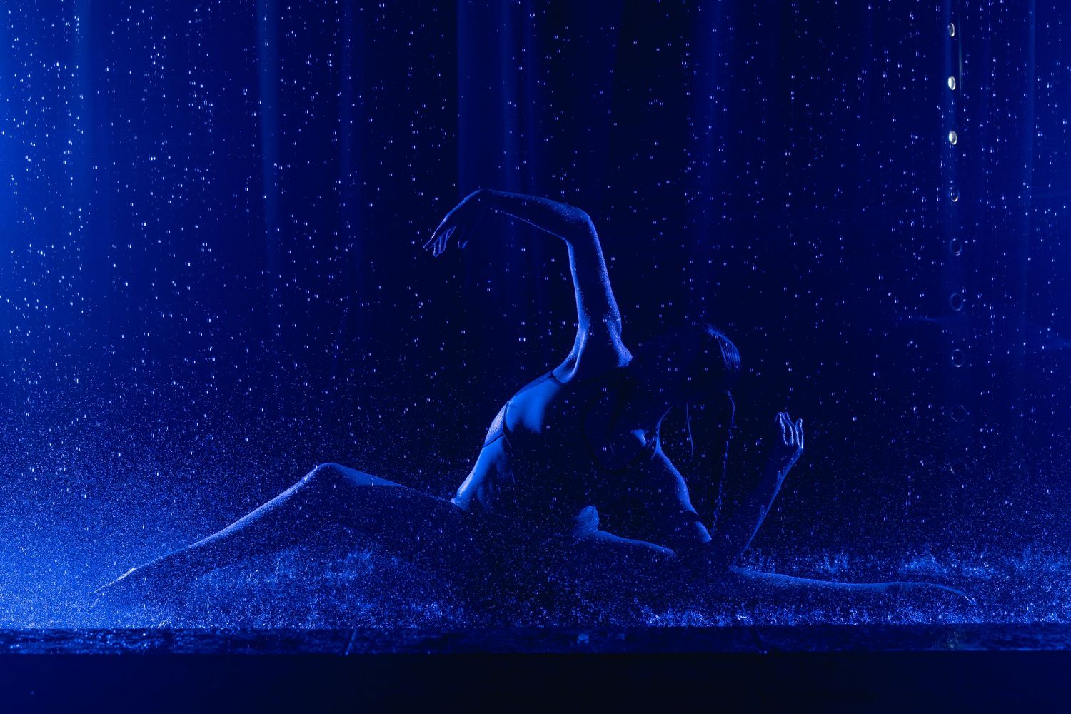 Two dancers perform an intimate silhouette routine under blue-lit falling water droplets on a dark stage.