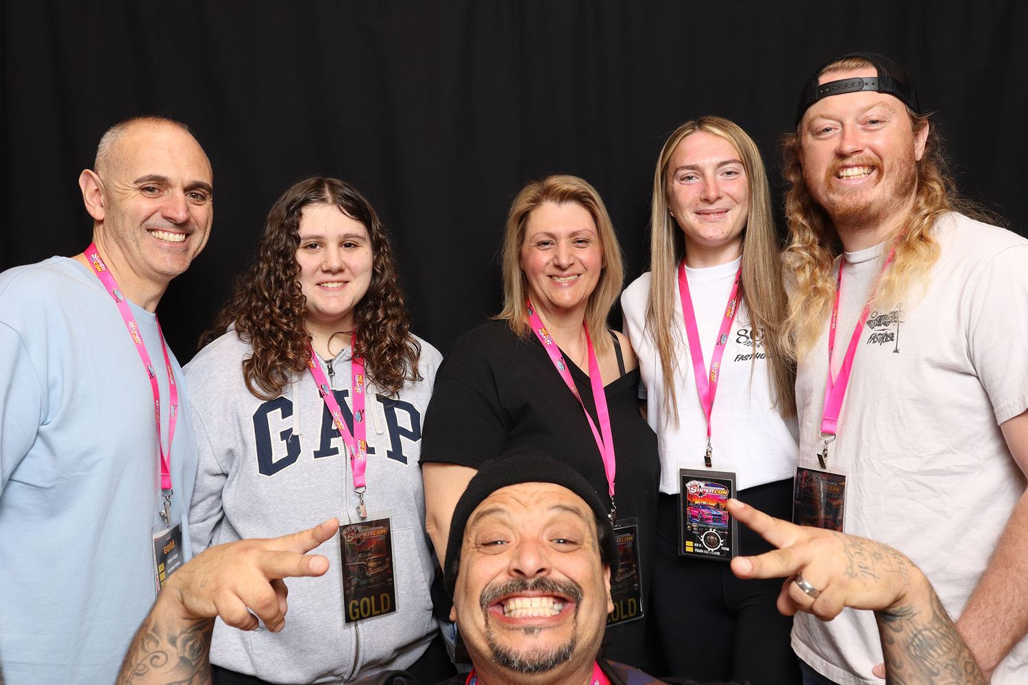 Group of six people smiling at a meet and greet event, wearing pink lanyards against a black backdrop.