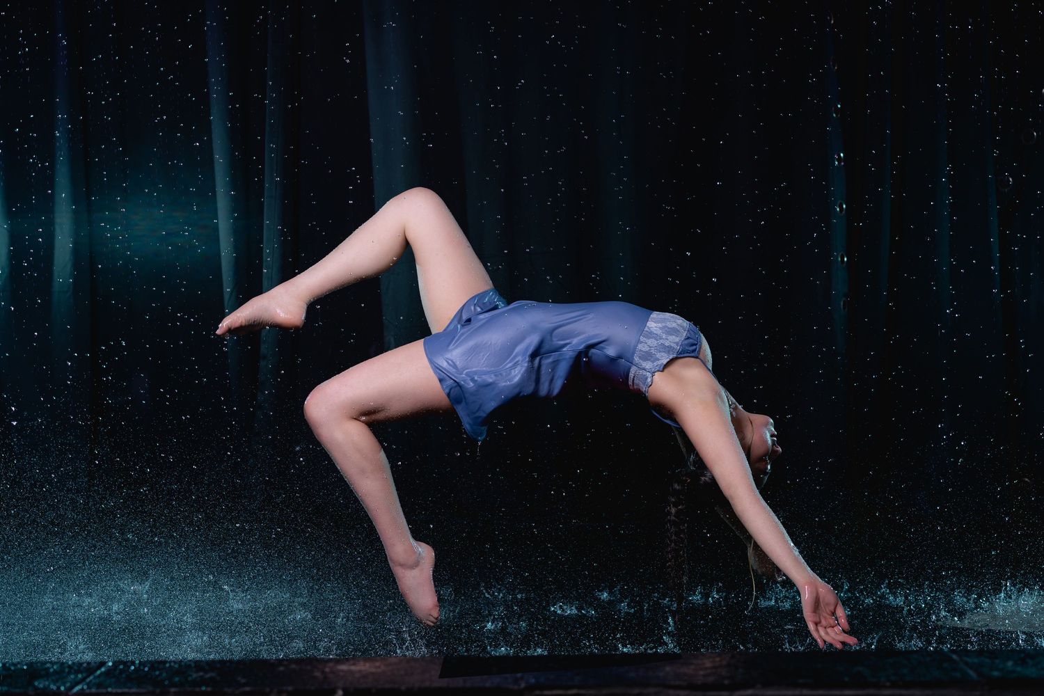Female dancer performing a dramatic backbend pose on stage with blue atmospheric lighting and rain effects.