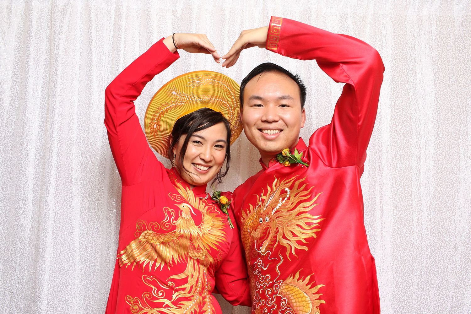 Vietnamese couple in red ao dai traditional wedding attire forming a heart shape with their arms, smiling at camera.