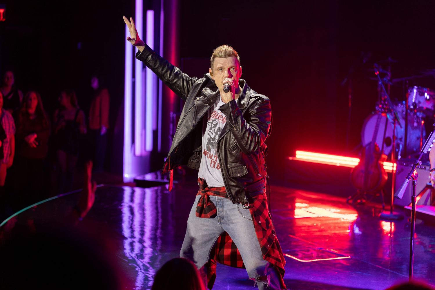 Male performer sings on stage wearing leather jacket under red neon concert lights.