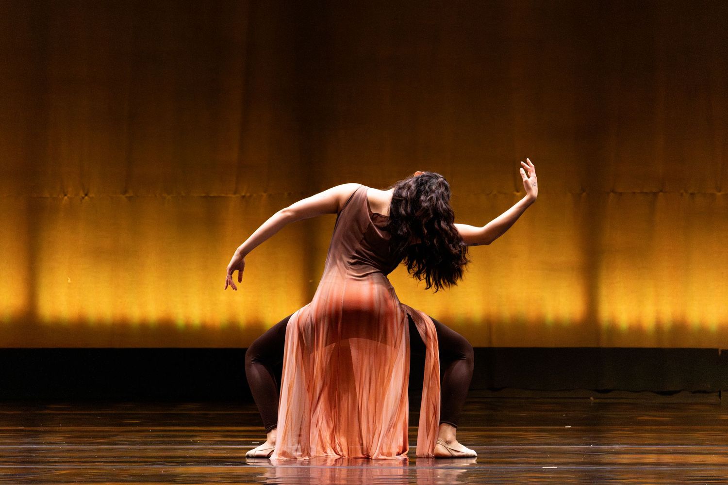 Graceful dancer in flowing peach gown performs expressive floor routine on warmly lit golden stage.